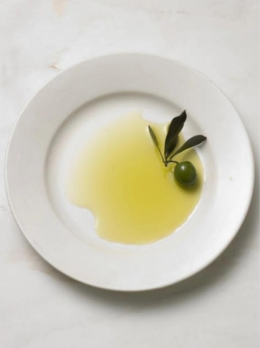 Extra virgin olive oil served on a white tasting plate with a fresh green olive and olive leaves.